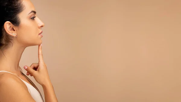 Neck lift, platysmaplasty. Side view of brunette hispanic millennial woman wearing top touching her chin and looking at copy space, showing youth-looking neck, isolated on beige background, panorama