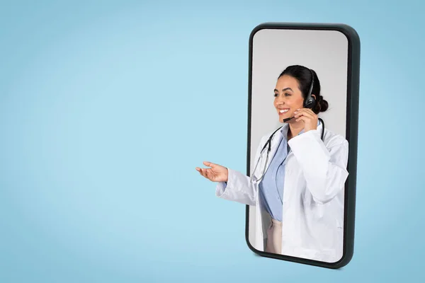 Happy cheerful latin woman doctor with headset in big cellphone screen gesturing and smiling, having online appointment, blue background, copy space