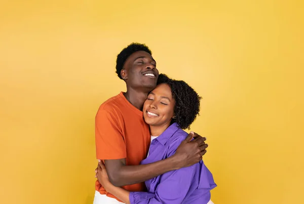 Happy millennial black guy hugging lady, enjoy love, isolated on yellow studio background, copy space. Romantic date together, free time, couple emotions, lifestyle, ad and offer