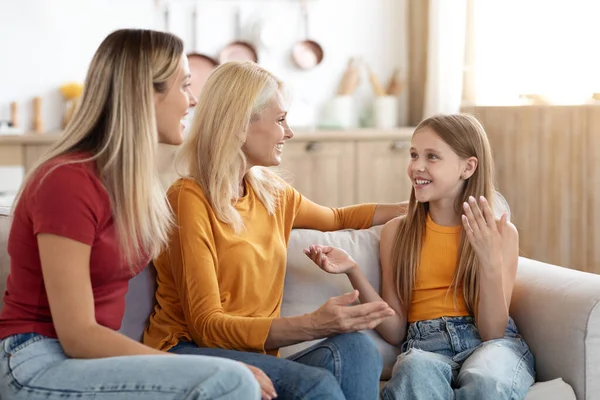 Cute preteen blonde girl sharing secrets with mom and granny at home, family three generations mother, daughter and grandmother sitting on couch in living room, have conversation, smiling