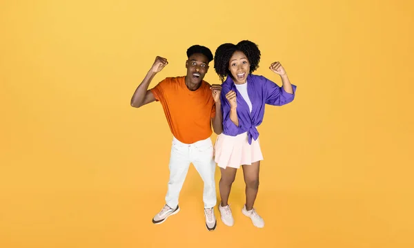 Happy millennial black lady and guy in casual celebrating victory, making success gesture, isolated on yellow studio background. Human emotions, surprise, sale, good news, lifestyle ad and offer