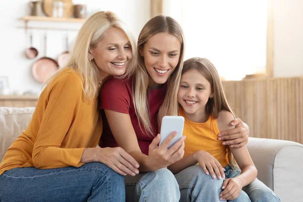 Happy caucasian family three generations mother, daughter and grandmother sitting on couch at home, using smartphone, shopping online, scrolling on Internet, copy space. Family offer, online deal