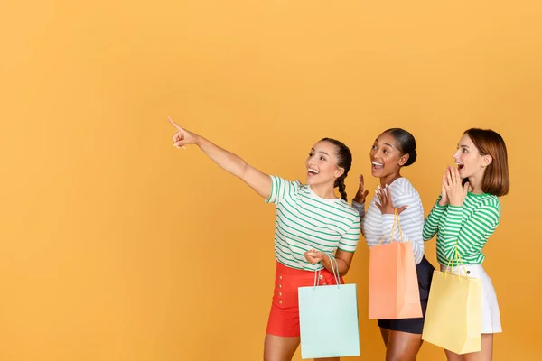 広告やテキストのための空白スペースを指すカラフルなショッピングバッグを運ぶカジュアルな衣装で幸せな多種多様な若い美しい女性 素晴らしい販売シーズンのオファーを示す オレンジ色の背景 — ストック写真