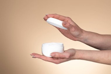 Olgun bir kadın yüz kremi ile kavanozu tutuyor ve açıyor, bej rengi stüdyo arka planının başında duruyor, yakın plan, kopyalama alanı, tasarım afişi