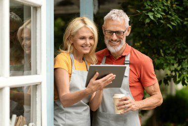Mutlu olgun çift kahve içiyor ve dijital tablet kullanıyor, kıdemli eşler gülümsüyor küçük işletme sahipleri muhasebe ve muhasebe için modern uygulama kullanıyorlar, kafelerinin terasında duruyorlar.