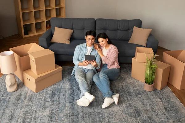 Real Estate Website. Happy Asian Young Couple Using Digital Tablet Sitting Among Carton Moving Boxes, Browsing Via Computer Searching House Apartment Rent Offers Online At Home
