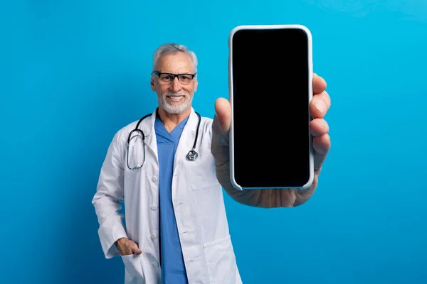 Telemedicine and Virtual Consultations. Handsome cheerful elderly doctor showing big smartphone with blank screen. Remote appointments, doc diagnose patients, provide medical advice, blue background
