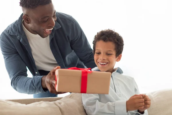 Loving african american dad giving gift box to little son, greeting boy and congratulating him on birthday at modern home interior. Family celebrating holiday, father gives present to kid