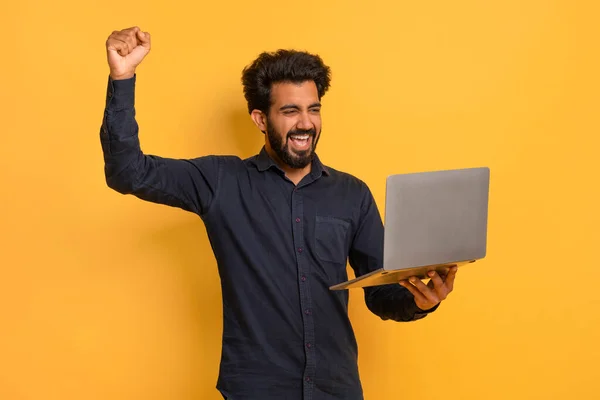 兴高采烈的印度年轻人手握手提电脑庆祝成功 兴奋的东方人高举断断续续的拳头 欢呼雀跃 欢呼胜利与好运 同时站在黄色的背景下 — 图库照片