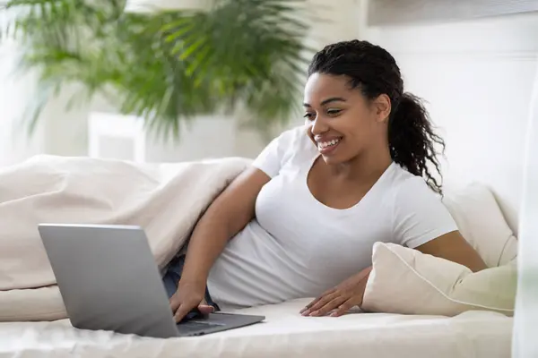 Domestic Leisure. Beautiful Young Black Woman Using Laptop While Lying In Bed, Happy African American Female Browsing Internet On Computer And Relaxing In Cozy Bedroom At Home, Copy Space