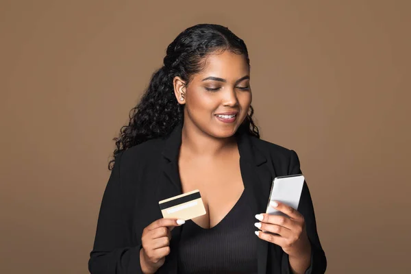 Financial offer. Cheerful brazilian plus size woman using cellphone and credit card, advertising bank service over brown studio background. Female customer posing with phone and bankcard