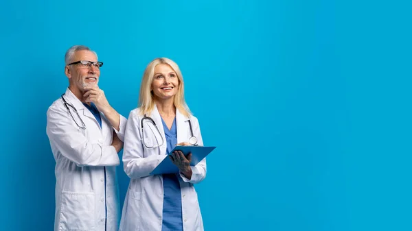 Healthcare, medical, modern clinic, hospital concept. Cheerful excited senior doctors man and woman in medical uniform looking at copy space, isolated on blue background, panorama