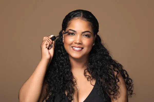 Beauty portrait of charming brazilian chubby woman applying mascara on her lashes over brown studio background, using organic makeup, putting on decorative cosmetics