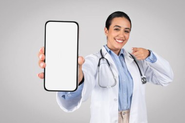 Boş cep telefonu ekranı, reklam kliniği web sitesi ya da tıbbi uygulama gösteren mutlu Latin kadın doktor gri stüdyo arka planında akıllı telefonu işaret ediyor.