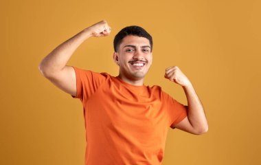 Biseps kaslarını gösteren güçlü Brezilyalı bir adam sarı stüdyo arka planında duruyor, turuncu tişört giyiyor ve kameraya gülümsüyor. Kuvvet kavramı