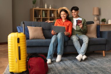 Mutlu Asyalı çift ellerinde pasaport, biletlerle dizüstü bilgisayar tutuyorlar, evlerinde uçuş ve otel rezervasyonu yapıyorlar, bavullu turistler tatil planlıyorlar, geziye çıkıyorlar.
