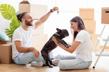 İpotek, insanlar ve gayrimenkul konsepti. Mutlu çok ırklı çift, Fransız köpeğini kucaklıyor, kutu kutu yerde oturuyor, anahtarı gösteriyor, taşınma gününün tadını çıkarıyor, mola veriyorlar.