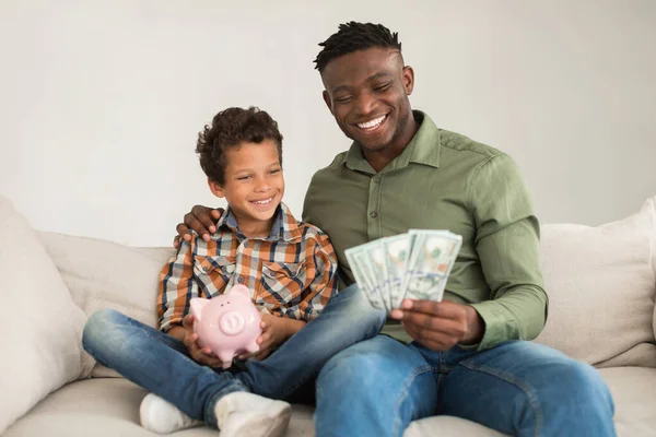 Happy Black Father And His Little Son Posing With Piggyback And Money Cash, Holding Dollar Banknotes Advertising Great Bank Offer, Sitting On Sofa At Home. Financial Investments Concept