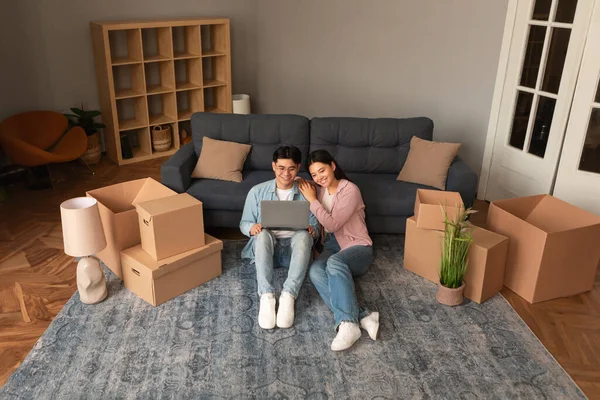Real Estate Website Ad. Japanese Spouses Using Laptop Computer Surfing Internet, Moving Choosing Property For Sale, Sitting Among Cardboard Boxes Indoors. New House Owners Relaxing With PC At Home