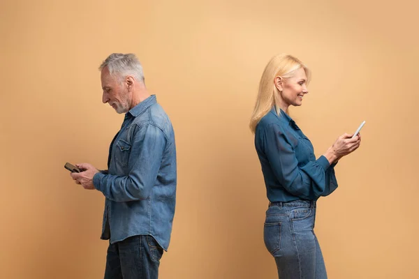 Gadget addiction, cheating in relationships, dishonesty in marriage. Mature man and woman standing back to back, using smartphones, isolated on beige background, empty space between them
