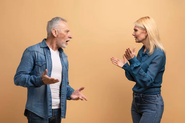 Unhappy marriage, crisis in relationships. Angry senior blonde woman wife have quarrel with her furious husband grey-haired man, elderly couple fighting over beige studio background