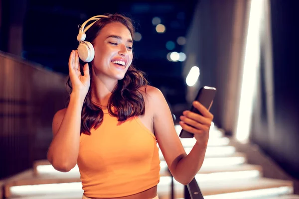 Excited woman enjoying favorite song during walk in the city at night, holding cellphone and smiling, free space. Music lover enjoying music outdoors