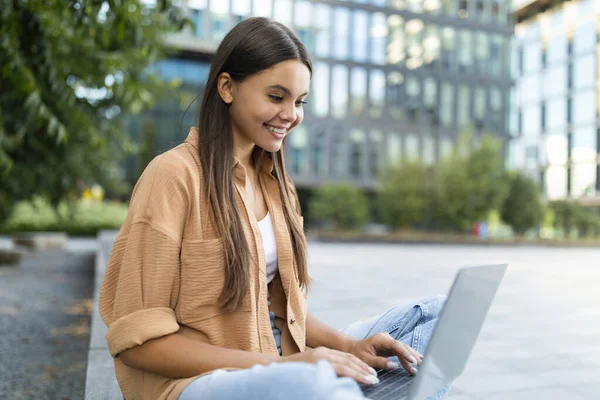 暑期在大学校园做作业时 留着长发的年轻女子坐在长椅上 用电脑笔记本 快乐的女性自由职业者上网工作 复制空间 — 图库照片