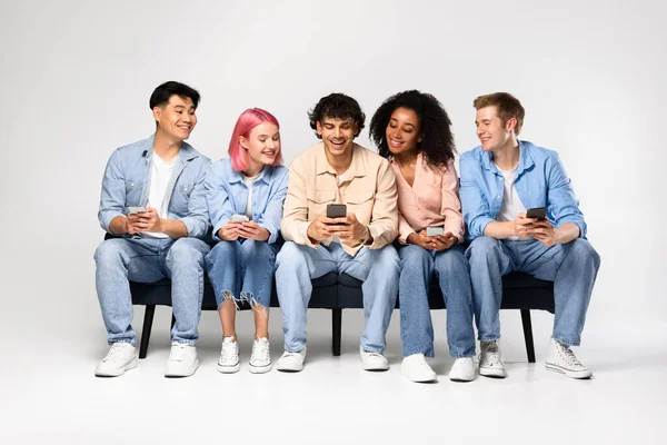 Happy multiracial friends sharing social media app news, sitting on sofa and holding phones, young people looking at guy smartphone, having fun together on white background