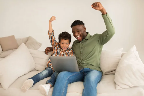 网上玩乐 快乐的黑人爸爸和小儿子一起使用笔记本电脑 快乐地挥动拳头 在家里一起庆祝电脑游戏的胜利 为父亲和孩子提供数码娱乐 — 图库照片