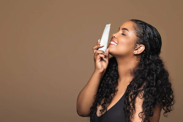 Happy brazilian chubby woman with perfect pure skin holding tube of cream near face, posing isolated on brown background, side view, copy space. Natural cosmetics, body and beauty care