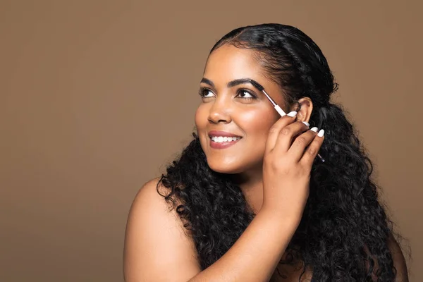 Eyebrow brushing process. Pretty brazilian body positive woman combing her eyebrows with brush tool, standing isolated on brown background, free space