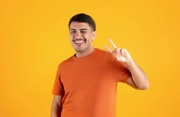 stock image Excited brazilian man making peace sign with hand and winking at camera, isolated on orange background, studio shot. Gesture, lifestyle, ad and offer, leisure time and fun