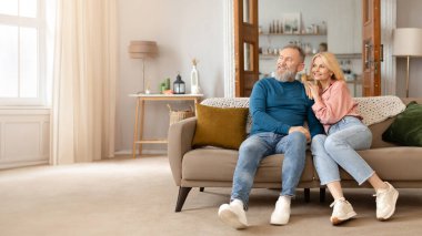 Mutlu Evli Son Sınıf Çifti koltukta dinleniyor, boş alana bakıyor, modern oturma odasında birlikte poz veriyor, yeni evin rahatlığının tadını çıkarıyor. Panorama