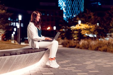 Dizüstü bilgisayarda çalışan, akşam parkında bankta oturan, modern kentsel arka plan, kenar manzara, serbest alan ile çalışan mutlu serbest çalışan kadın.