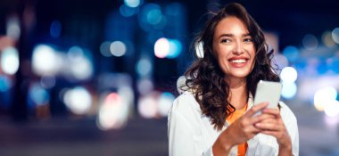 Cep telefonu tutan mutlu genç bayanın portresi, kenara bakıyor ve gülümsüyor, akşamleyin şehir caddesinde duruyor, arka planda neon ışıkları, boş alanı olan panorama.