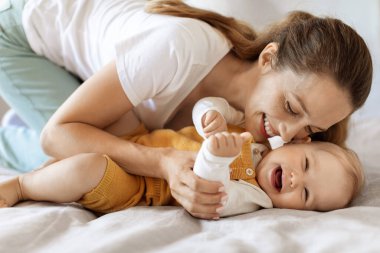 Evde yatakta kaynaşan mutlu anne ve bebeğin portresi. Güzel anne onu okşuyor ve öpüyor. Gülüşü çok tatlı, küçük oğluna gülüyor. Annelik, çocuk bakımı kavramı