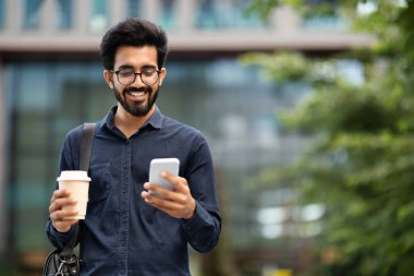 Pozitif neşeli genç Hintli adam şık günlük kıyafetler ve gözlük takıyor, çanta çalışanını ofise götürüyor, kahve içiyor, akıllı telefon kullanıyor ve gülümsüyor, fotokopi çekiyor.