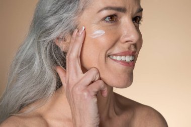 Sağlık ve güzellik. Mutlu Kafkas kıdemli kadın nemlendirici yüz, yanak kremi sürüyor ve gülümsüyor, bej arka planda poz veriyor, stüdyo çekimi