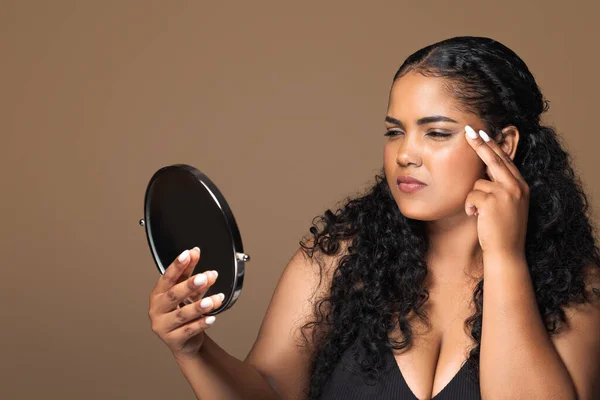 Concerned brazilian woman holding mirror and looking at wrinkles near her eyes, examining fine lines on face while making daily beauty routine, brown background, free space