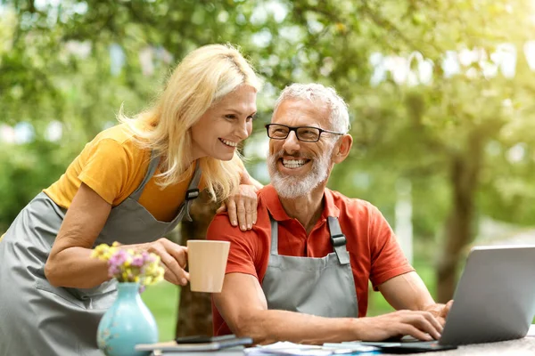 Modern Retirement Lifestyle. Happy senior couple using laptop outdoors at their terrace, smiling mature spouses in aprons browsing internet together, shopping online, spending weekend in countryside