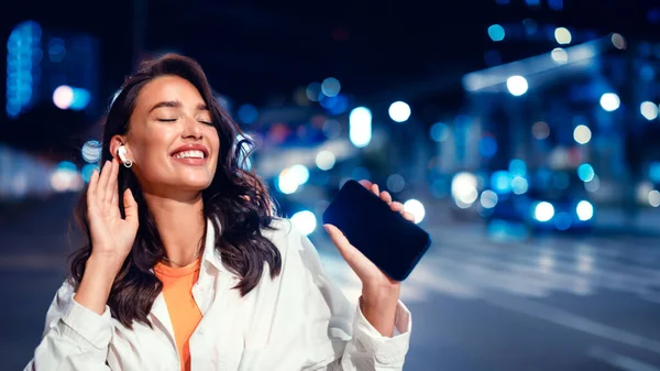 Joyful lady enjoying favourite music in earphones holding smartphone, walking at night on city street with neon illumination on background, panorama with free space