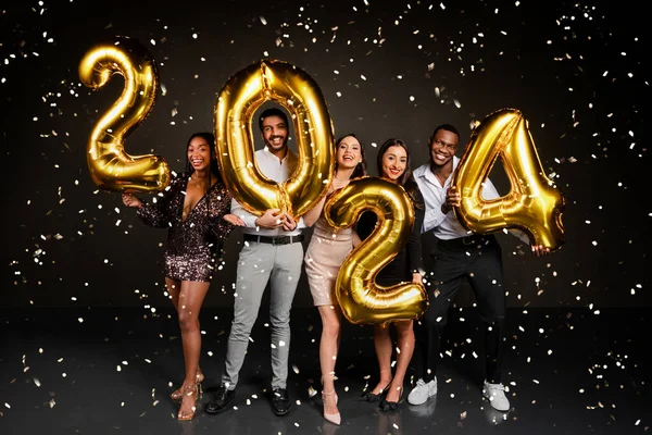 Excited beautiful multicultural friends men and women in nice outfits celebrating New Year 2024 together, holding golden number balloons and posing over black background, enjoying falling confetti