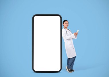 Gülümseyen Asyalı güzel bayan doktor beyaz ekranlı büyük boş akıllı telefonun yanında duruyor, üniformalı bir kadın elinde cep telefonu, hastalarla sohbet ediyor, mavi arka plan, model.