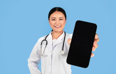 Asyalı Doktor Kadın Üniformalı Kadın, Siyah Ekranda Siyah Ekran 'la Büyük Boş Akıllı Telefon Gösterirken, Mavi Arkaplanda, Tıbbî Reklam İçin Kopya Alanı Gösteren Kadın Doktor,