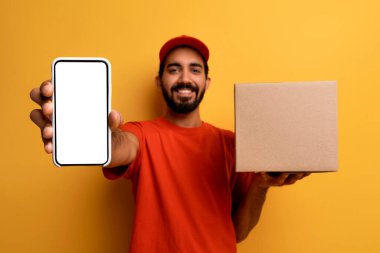 Mutlu Hintli kurye cep telefonunu boş ekranla gösteriyor ve elinde karton kutu, yakışıklı genç doğulu reklamcı teslimat servisi, sarı stüdyo arka planında poz verme, model...