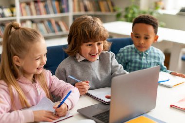 Üç farklı sınıf arkadaşı dizüstü bilgisayarın önünde oturuyor, ders sırasında online videolar izliyor, ekrana bakıyor ve gülümsüyor, sınıfta oturuyor. Eğitim kavramı için modern teknolojiler