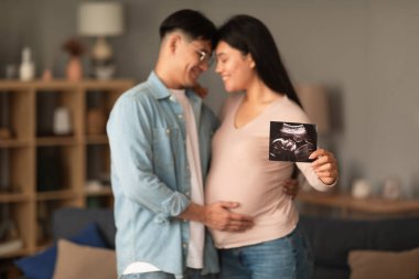 Hamilelik, bebek beklentisi. Asyalı çiftin kucaklaşması evde ultrason embriyo resim fotoğrafı gösteriyor, seçici odaklanma. Koca hamile kadının karnına dokunuyor ve çocuğunu bekliyor. Sığ derinlik