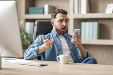 Ofiste otururken akıllı telefondan mesaj okuyan genç iş adamının portresi, cep telefonu ekranına bakan, kötü haberlere duygusal tepki veren Millennial Man