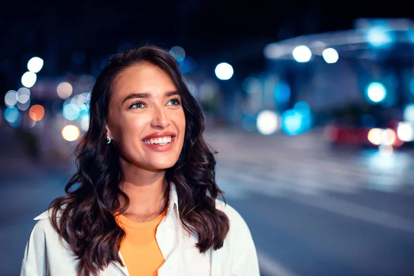 Excited caucasian woman walking on urban city street at night looking aside and smiling, bokeh and neon city lights on background, copy space