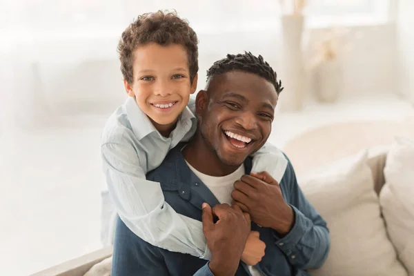 Daddy And Son Bond. Happy Little Boy Hugging His Black Father From Back, Posing Together And Expressing Positive Emotions At Home. Happy Family And Fatherhood Moments Concept